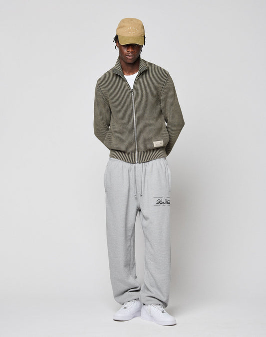 A person in a beige cap, LFDY Knit Jacket by LIVE FAST DIE YOUNG, light gray sweatpants, and white sneakers stands against a plain light background with hands in pockets, looking slightly downward.