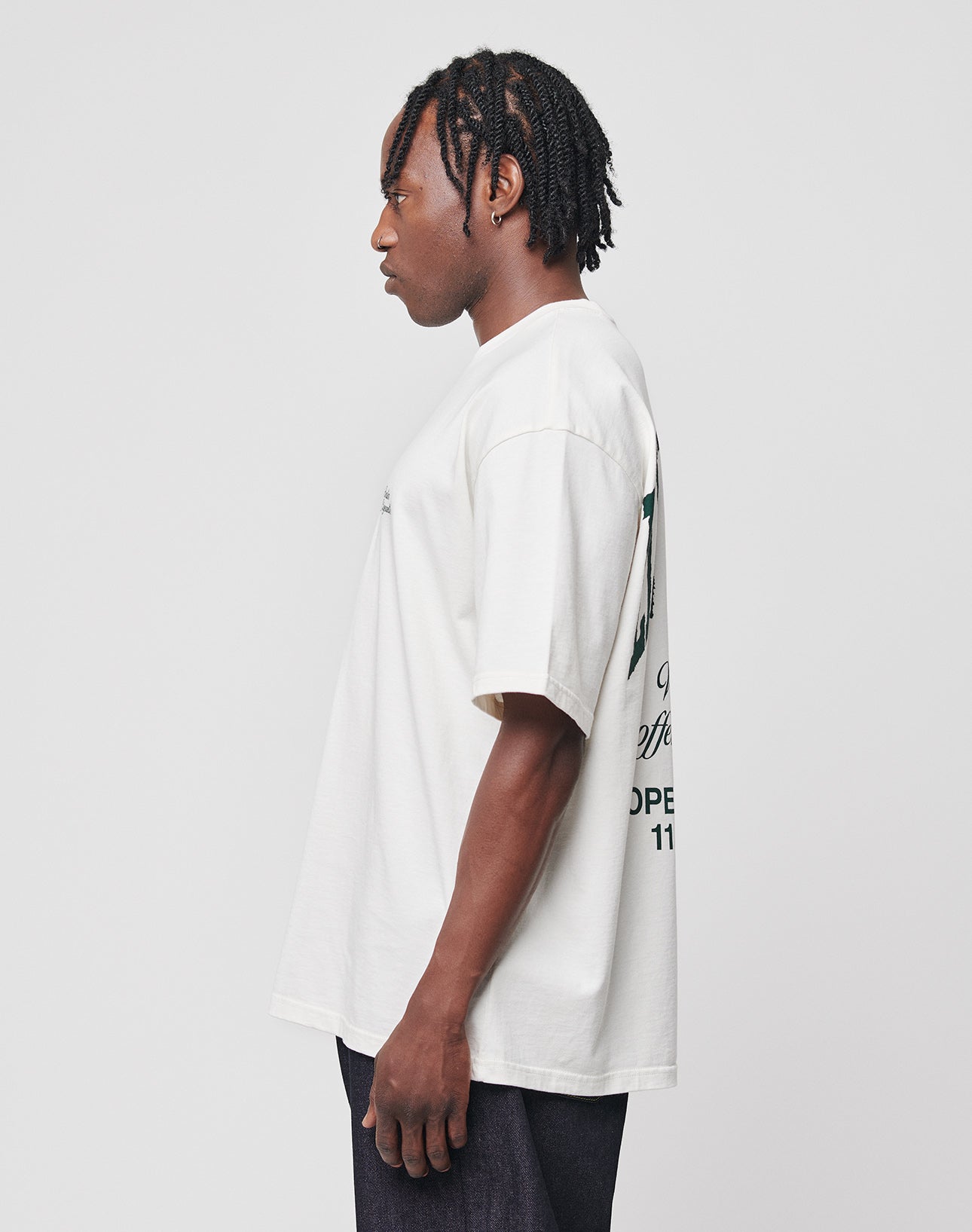 A man with short, braided hair and dark skin stands in profile against a light gray background, wearing the LIVE FAST DIE YOUNG Live Fast Coffee Tee—an oversize fit, 100% cotton white T-shirt with back graphics—in Berlin Café style.