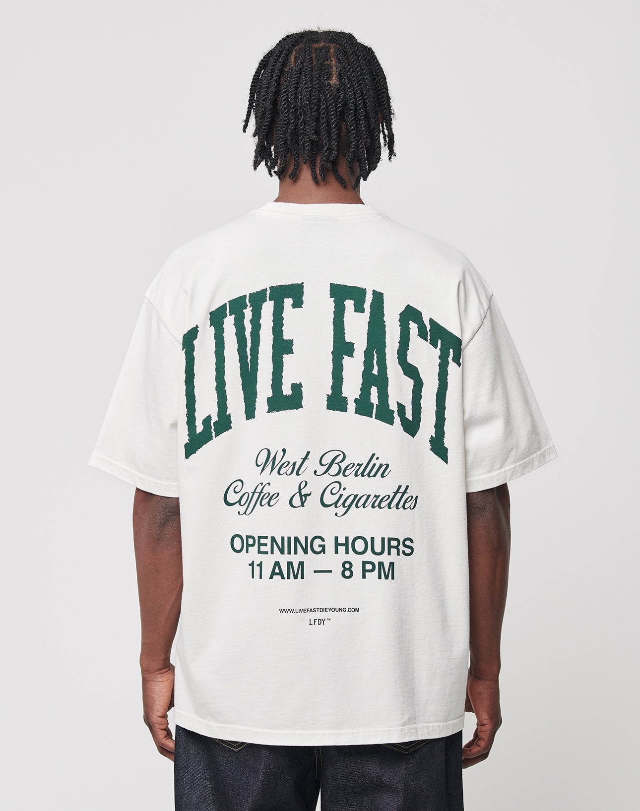 A person faces away from the camera, wearing the LIVE FAST DIE YOUNG Live Fast Coffee Tee—an oversize fit white T-shirt made of 100% cotton with green and black back text: “LIVE FAST, LFDY Berlin Café & Cigarettes, OPENING HOURS 11 AM – 8 PM.”. | model