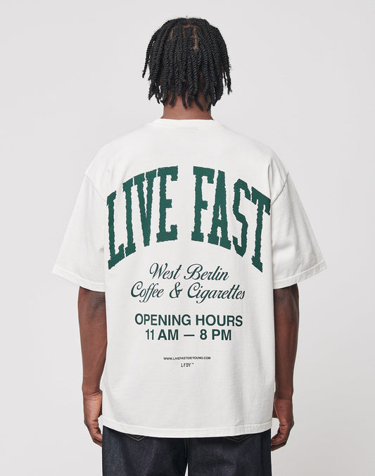 A person faces away from the camera, wearing the LIVE FAST DIE YOUNG Live Fast Coffee Tee—an oversize fit white T-shirt made of 100% cotton with green and black back text: “LIVE FAST, LFDY Berlin Café & Cigarettes, OPENING HOURS 11 AM – 8 PM.”. | model