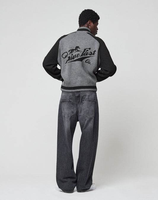 A person stands facing away in a LIVE FAST DIE YOUNG Boxy Knit Zip Jacket with gray and black colors, an intarsia-knit horse graphic on the back, loose dark gray jeans, and white shoes against a plain light background.