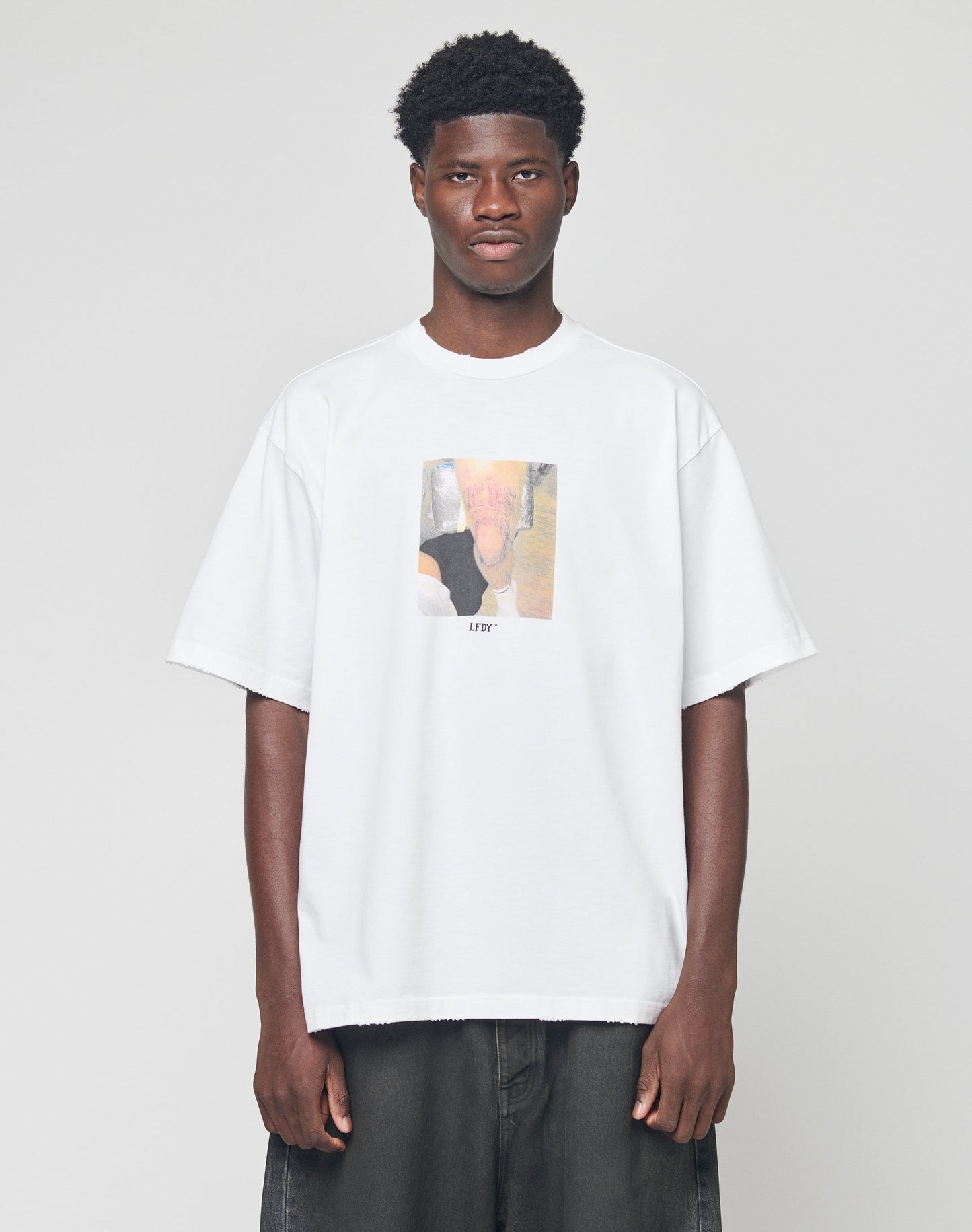 A man stands against a plain background wearing the LFDY Sharkhunter Tee by LIVE FAST DIE YOUNG, an oversize white 100% Baumwolle T-shirt featuring a blurry rectangle graphic and LFYT text on the front, paired with dark baggy jeans.