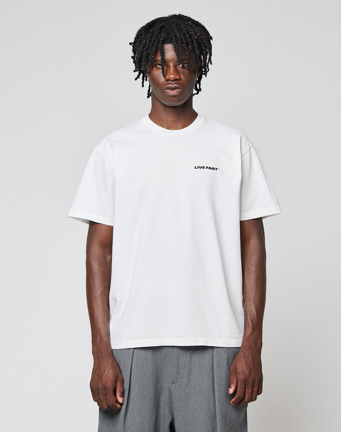 A person with short dreadlocks wears the LIVE FAST DIE YOUNG Live Fast Plain Tee, a regular fit white t-shirt with LIVE FAST. in small black letters on the left chest, made from 100% Baumwolle, paired with loose gray pants against a light background.