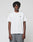 A person with short dreadlocks wears the LIVE FAST DIE YOUNG Live Fast Plain Tee, a regular fit white t-shirt with LIVE FAST. in small black letters on the left chest, made from 100% Baumwolle, paired with loose gray pants against a light background.
