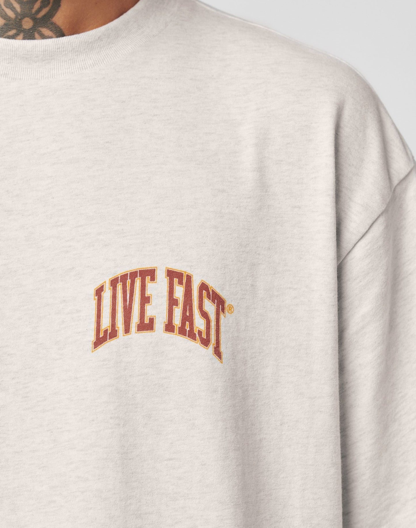 Close-up of a person in the LIVE FAST DIE YOUNG Live Fast Amsterdam Tee, an oversize light gray T-shirt featuring LIVE FAST in bold red and yellow on the chest. A neck tattoo adds a vintage vibe.
