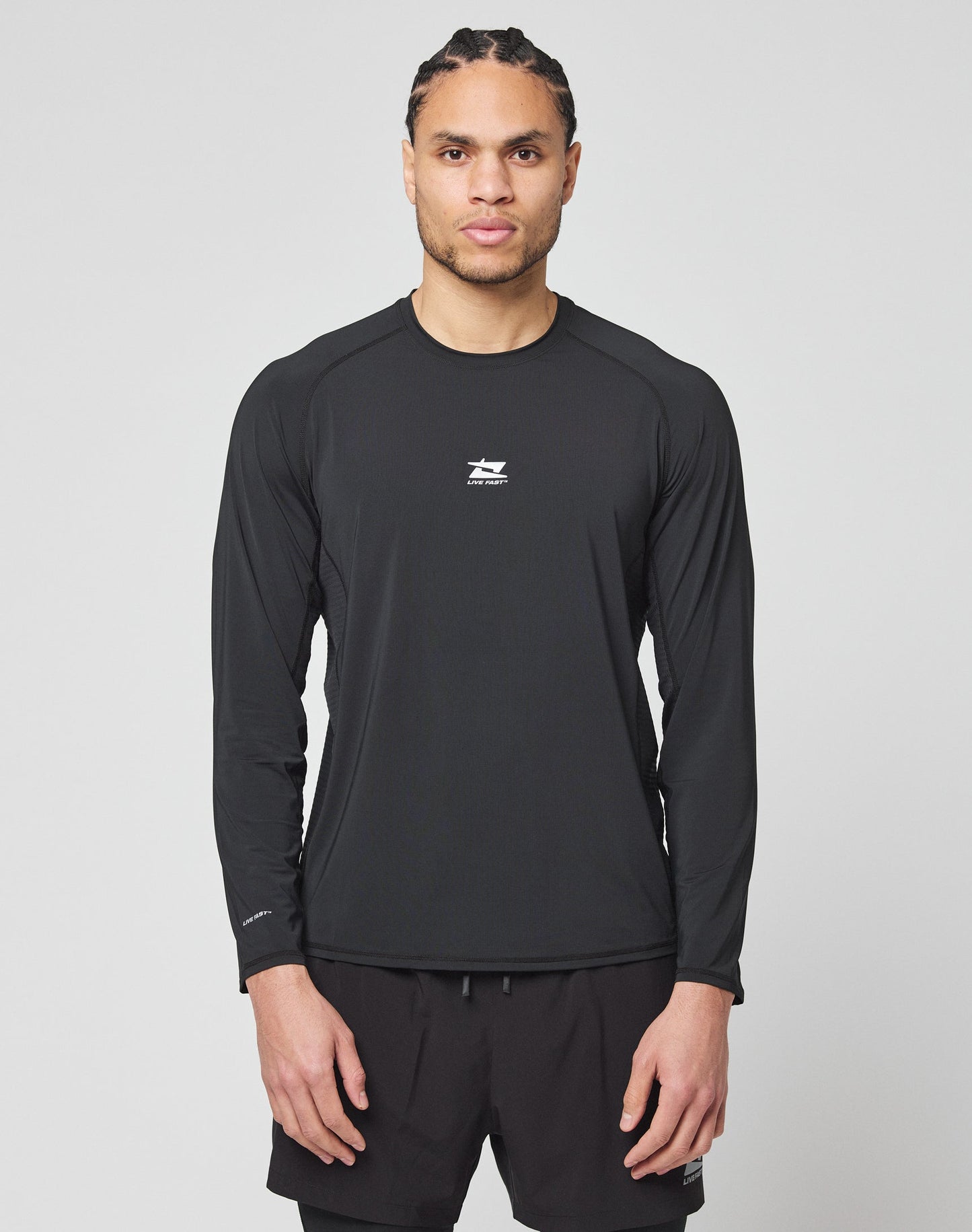 A man with braided hair wears the LIVE FAST DIE YOUNG Live Fast Performance Longsleeve in black and matching black shorts, standing facing forward against a plain light gray background. | model