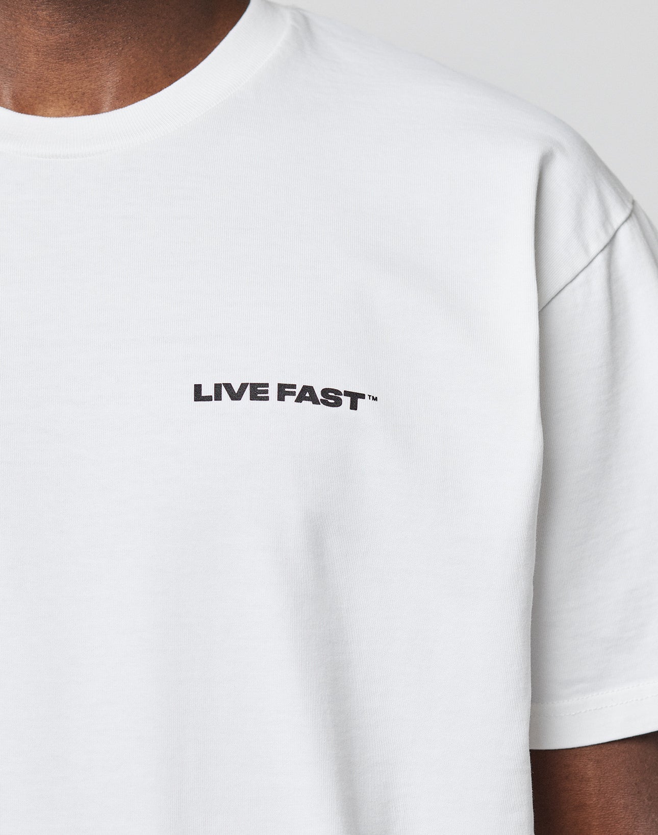 A person wears the LIVE FAST DIE YOUNG Live Fast Plain Tee, a 100% cotton white T-shirt with LIVE FAST printed in black on the left chest. Only the upper torso is shown.