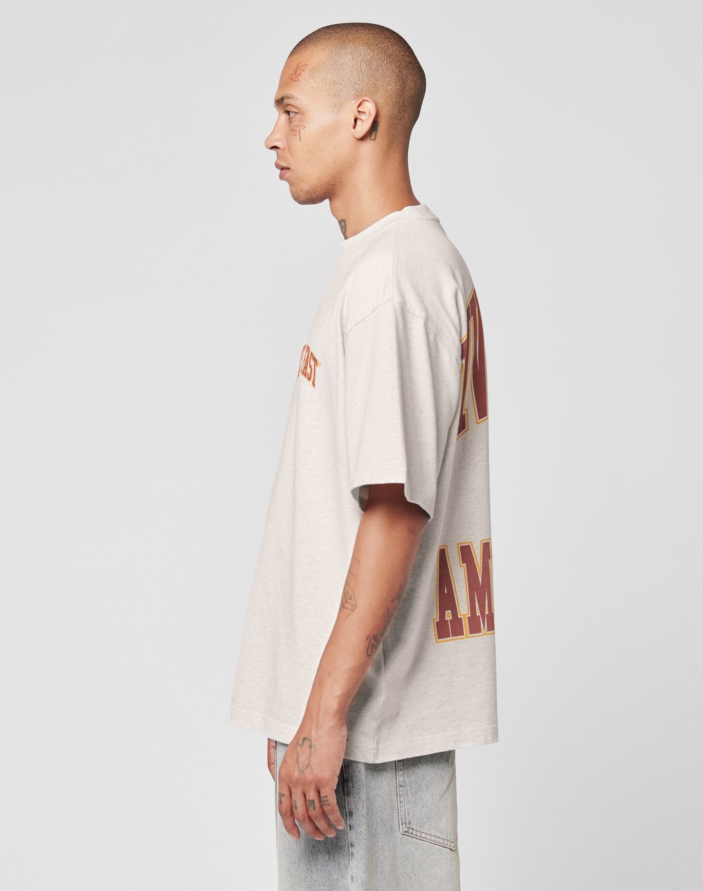 A person with a shaved head and tattoos stands in profile, wearing the LIVE FAST DIE YOUNG Live Fast Amsterdam Tee in light gray oversize fit with red and yellow lettering, paired with light blue jeans against a plain white background.