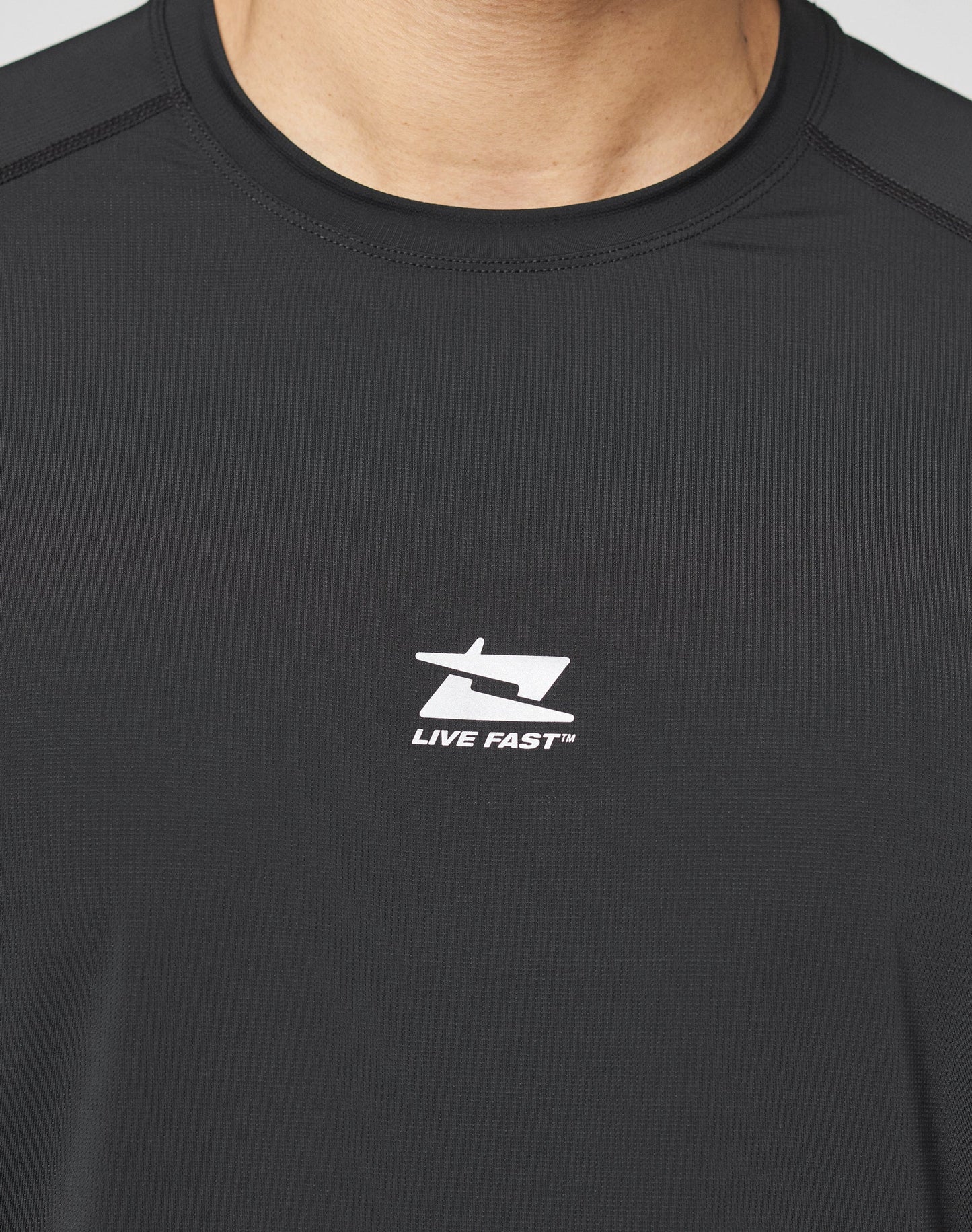 Close-up of a person wearing the black Live Fast Performance Longsleeve by LIVE FAST DIE YOUNG, featuring a white stylized logo and LIVE FAST™ as a reflective front print. The persons lower face and neck are partially visible.
