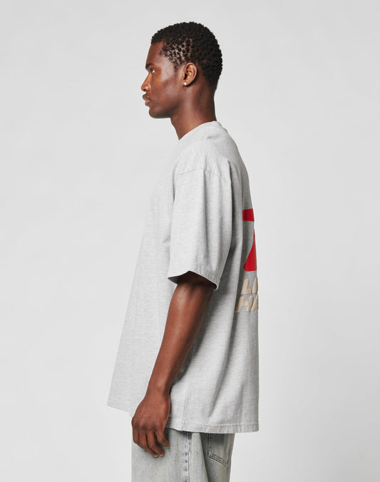 Someone in profile wears the oversize LFDY Hardwear Tee by LIVE FAST DIE YOUNG in light gray, featuring a red and white back design, paired with light blue jeans against a plain light background.