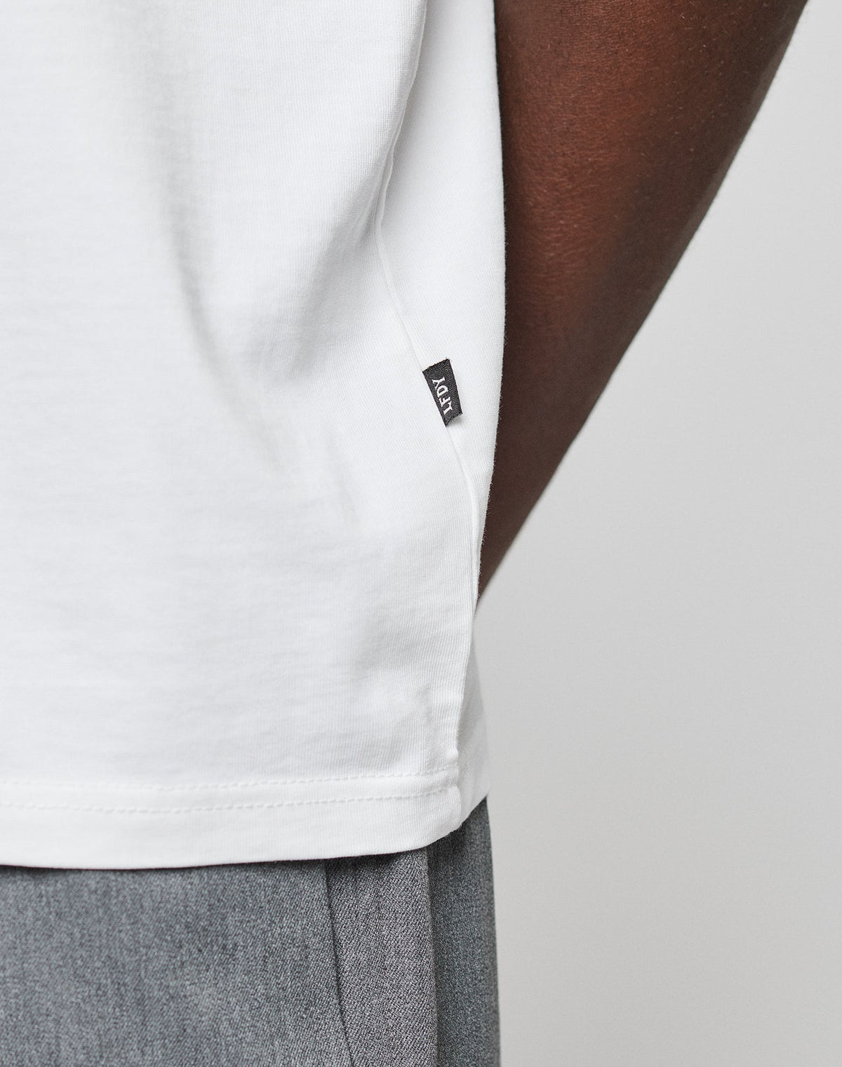 Close-up of a person wearing the white Live Fast Plain Tee by LIVE FAST DIE YOUNG, crafted from 100% Baumwolle, paired with gray pants. A small black brand tag is sewn into the side seam. Set against a plain, light background.