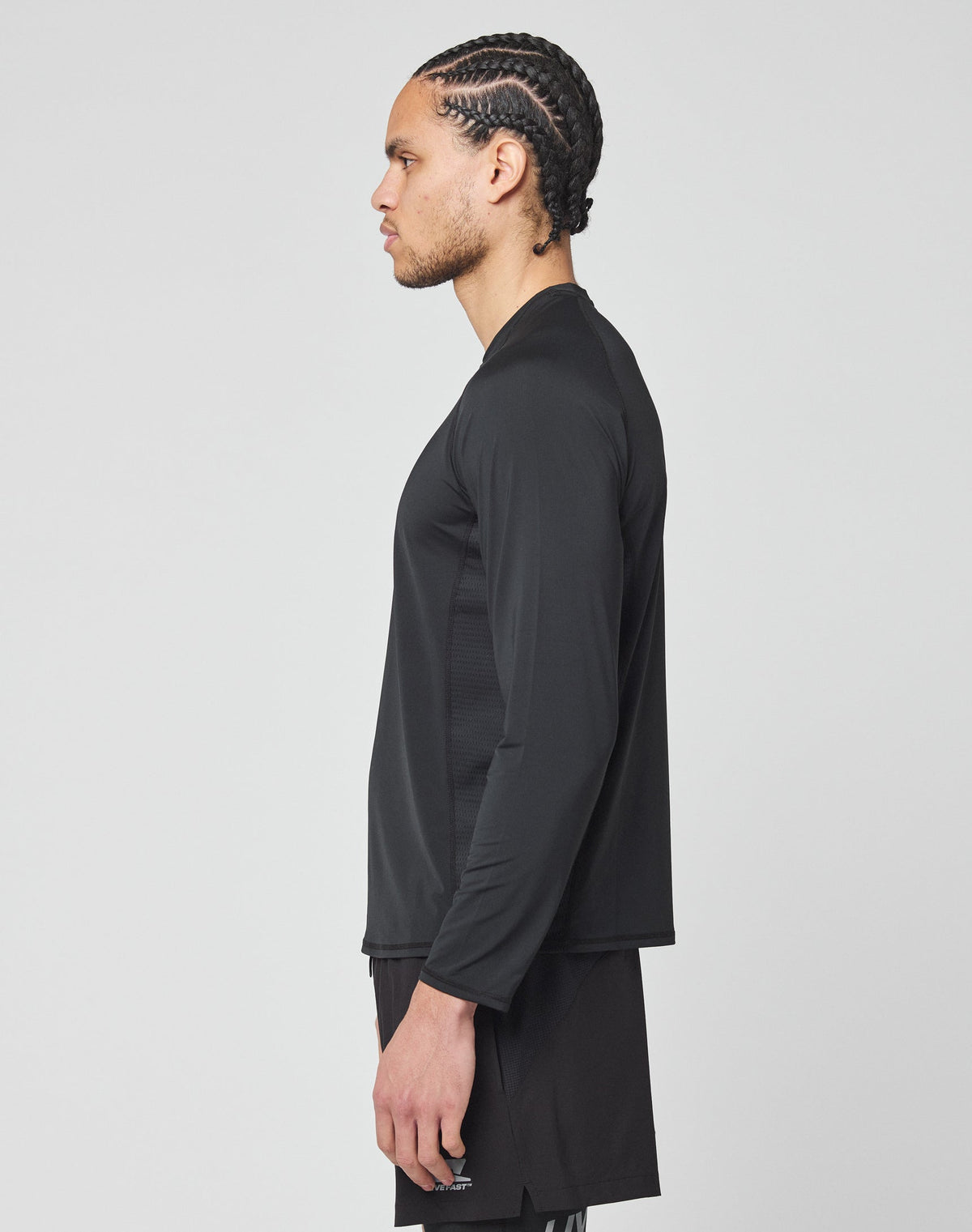 A man with braided hair stands in profile, wearing the LIVE FAST DIE YOUNG Live Fast Performance Longsleeve in slim fit and black shorts with a mesh insert, set against a plain light gray background.