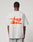 A person wears the LIVE FAST DIE YOUNG LFDY Hardwear Tee, a light gray oversized fit t-shirt featuring an orange abstract graphic and LIVE FAST™ HARDWEAR on the back, standing before a plain light background.