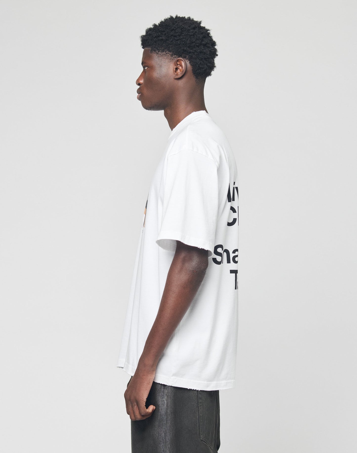 A young man with short curly hair stands in profile against a plain background, wearing the LIVE FAST DIE YOUNG LFDY Sharkhunter Tee—an oversized white t-shirt made from 100% Baumwolle with black text on the back—and dark pants.