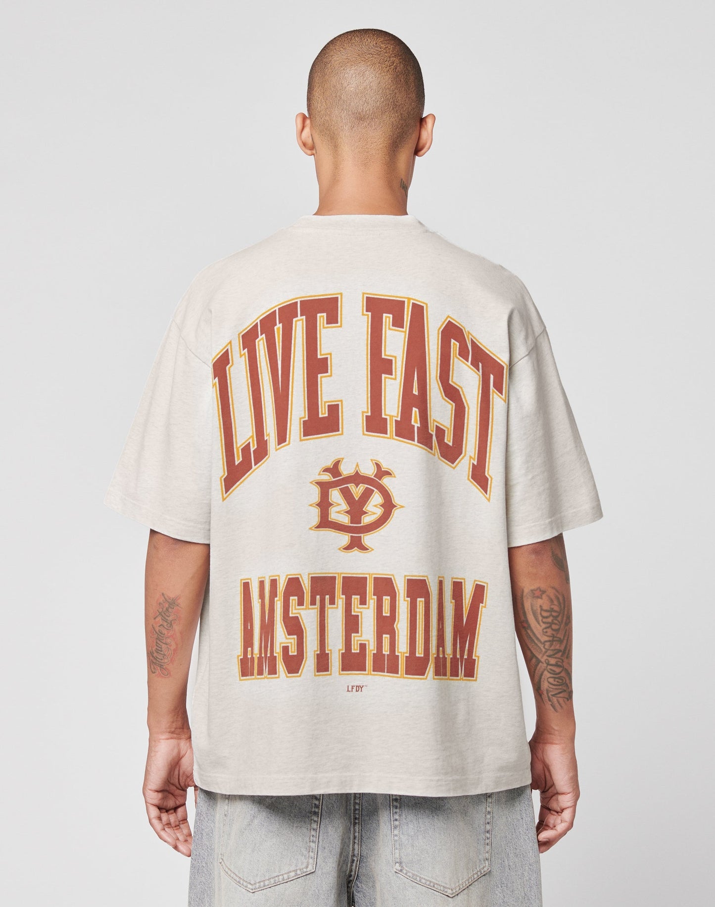 A person with a shaved head, seen from behind, wears the beige Live Fast Amsterdam Tee by LIVE FAST DIE YOUNG, featuring bold red and yellow LIVE FAST and AMSTERDAM text with a circular logo in between.