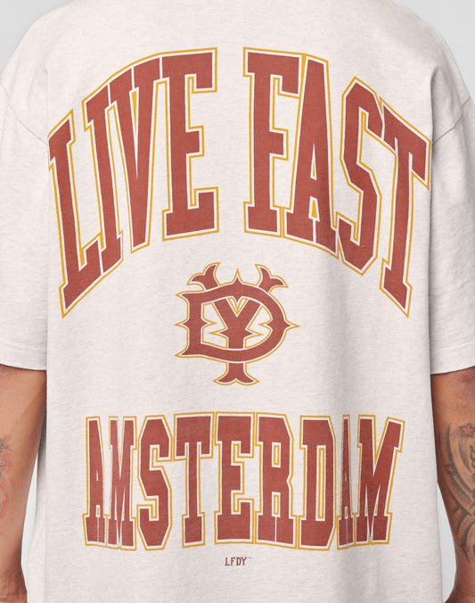 A person wears the Live Fast Amsterdam Tee by LIVE FAST DIE YOUNG—an oversized light gray cotton shirt with bold red and yellow LIVE FAST at the top, AMSTERDAM at the bottom, and a vintage-inspired logo on the back.