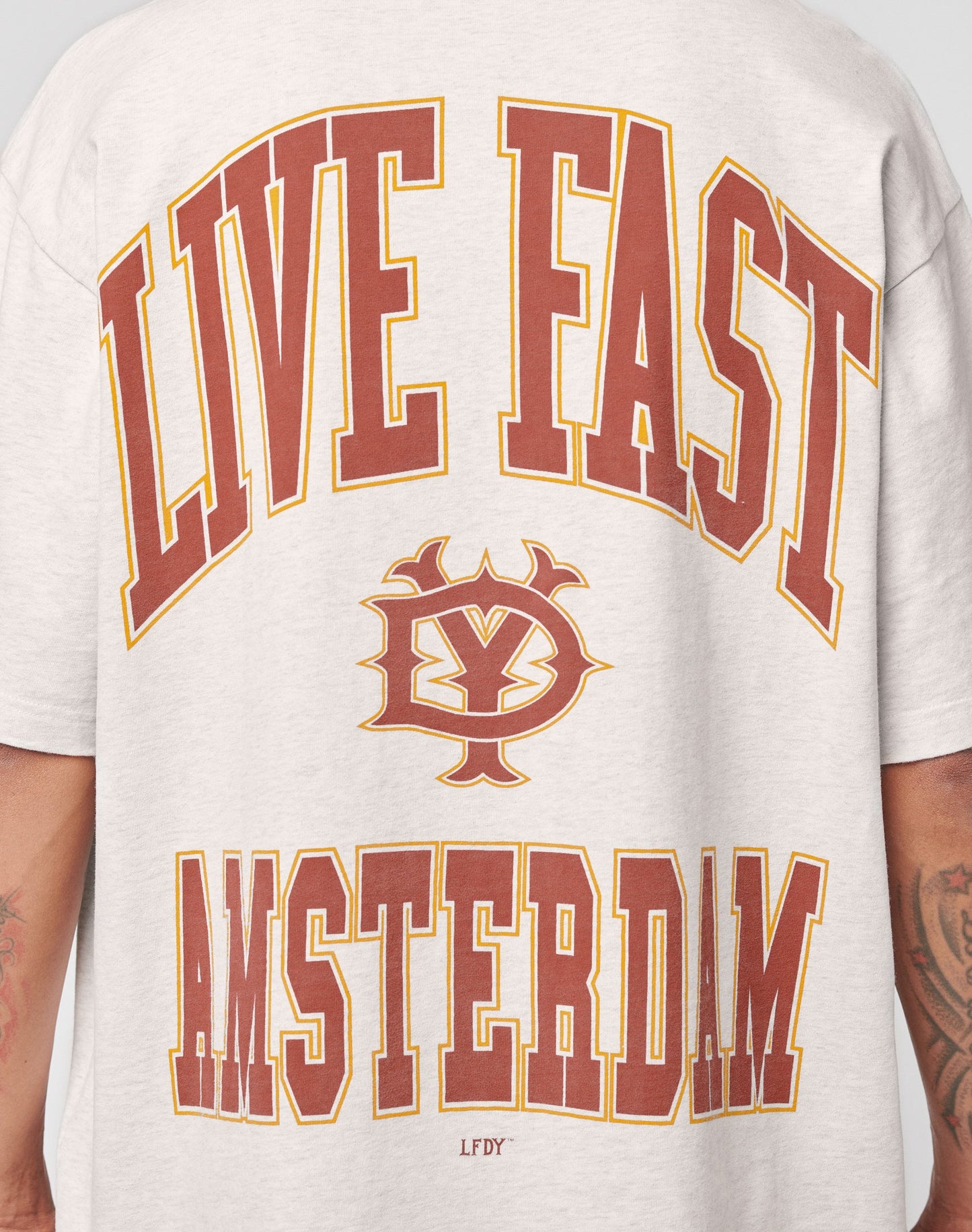 A person wears the Live Fast Amsterdam Tee by LIVE FAST DIE YOUNG—an oversized light gray cotton shirt with bold red and yellow LIVE FAST at the top, AMSTERDAM at the bottom, and a vintage-inspired logo on the back.