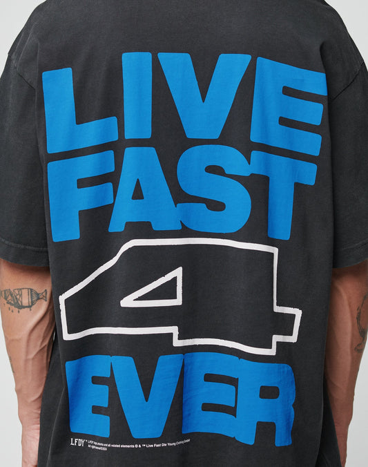 A person wears the LFDY 4 Ever Tee by LIVE FAST DIE YOUNG, an oversized black cotton T-shirt with a bold blue and white LIVE FAST 4 EVER backprint, styled over tattooed arms with a long-sleeve layer underneath.