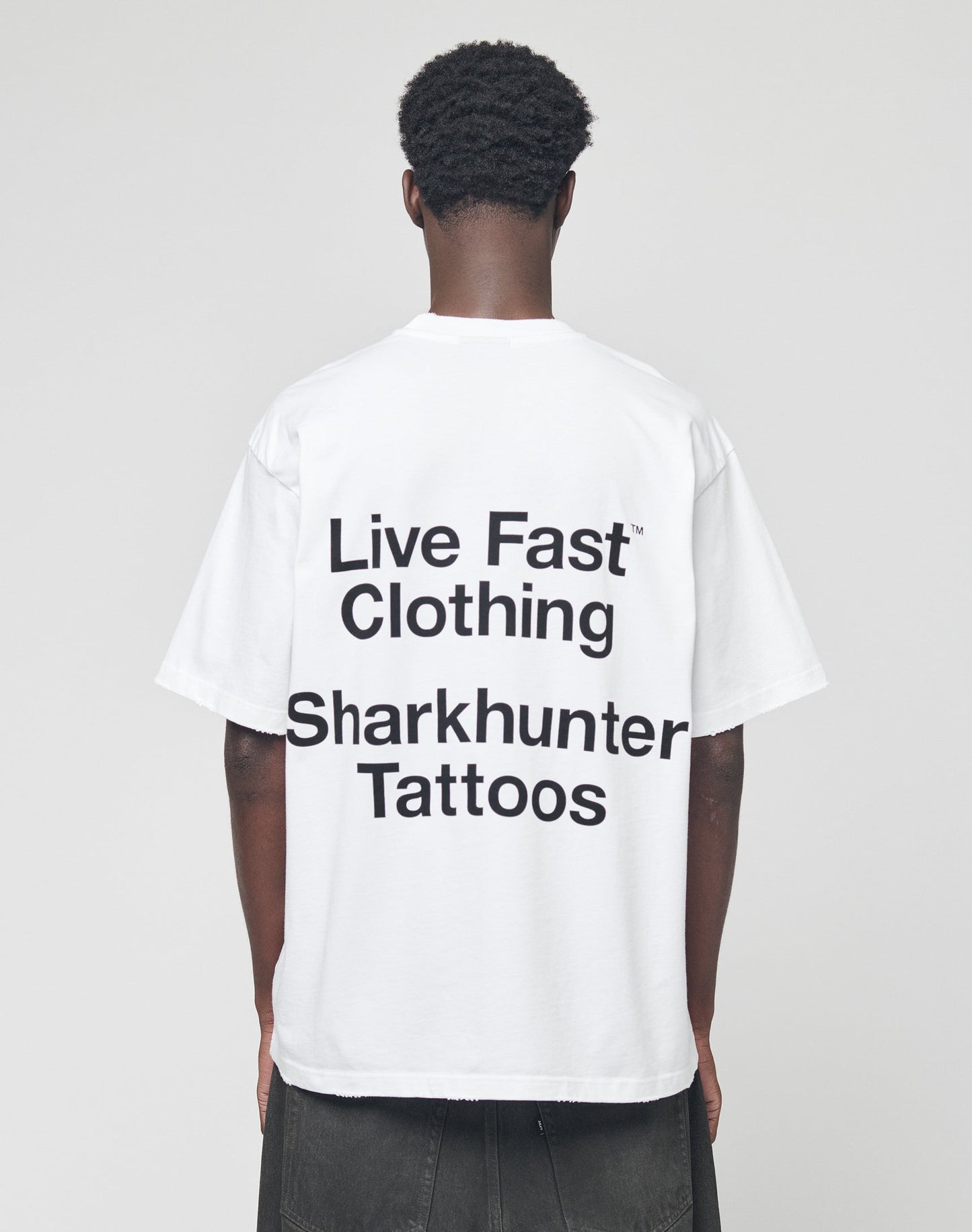 A person stands facing away, wearing the LFDY Sharkhunter Tee by LIVE FAST DIE YOUNG—an oversize white shirt with black Sharkhunter Tattoos text. Made from 100% Baumwolle, it’s set against a plain light gray background.