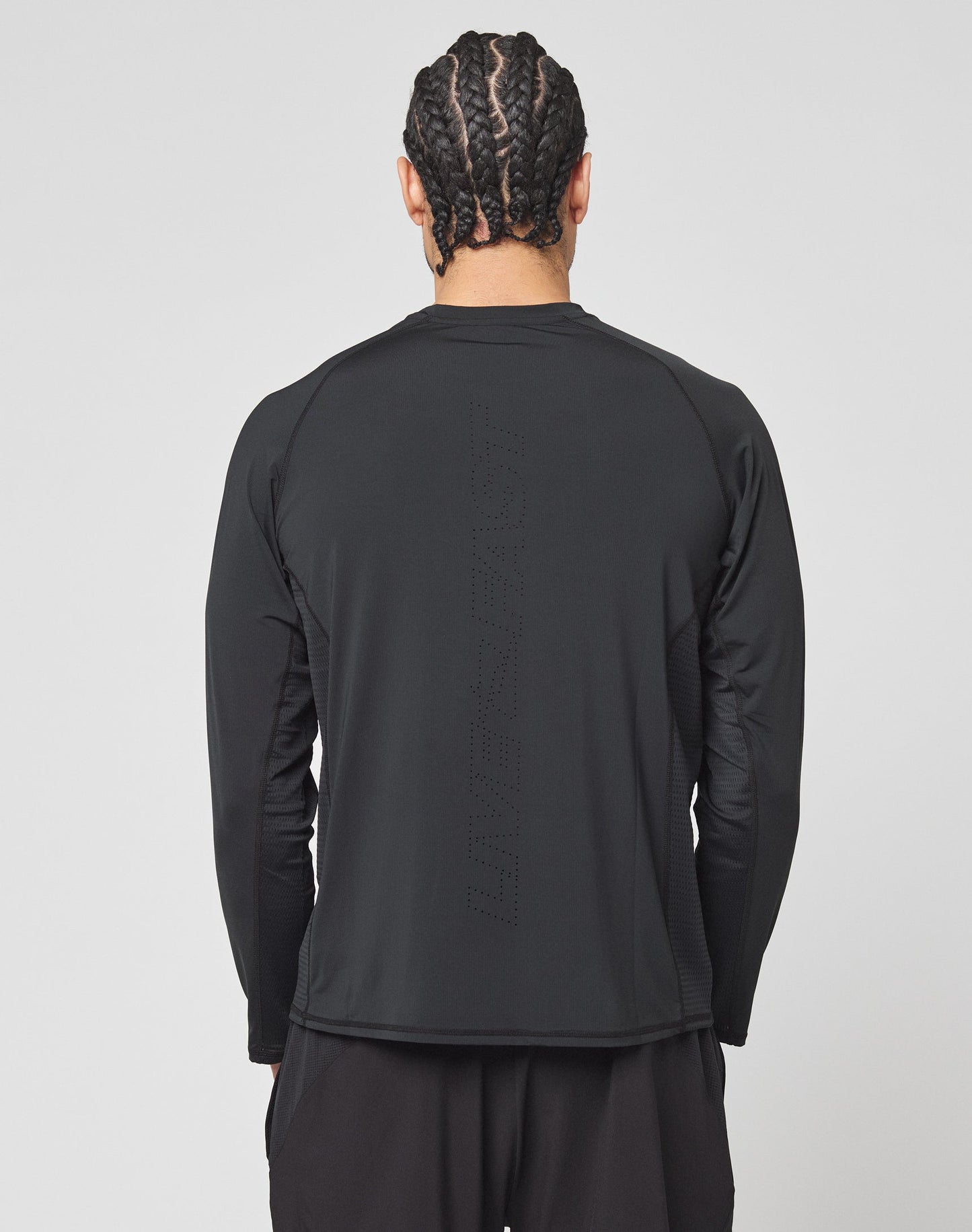 A person with braided hair is seen from behind in a LIVE FAST DIE YOUNG Live Fast Performance Longsleeve—a slim fit black athletic shirt with mesh insert and subtle perforated vertical text. The background is plain and light-colored.