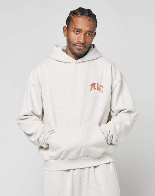 A man with braided hair wears the Live Fast Amsterdam Hooded sweatshirt and matching pants by LIVE FAST DIE YOUNG, in oversize fit light beige 100% Baumwolle, featuring a red LIVE FAST chest print. He stands hands in pockets against a plain background.