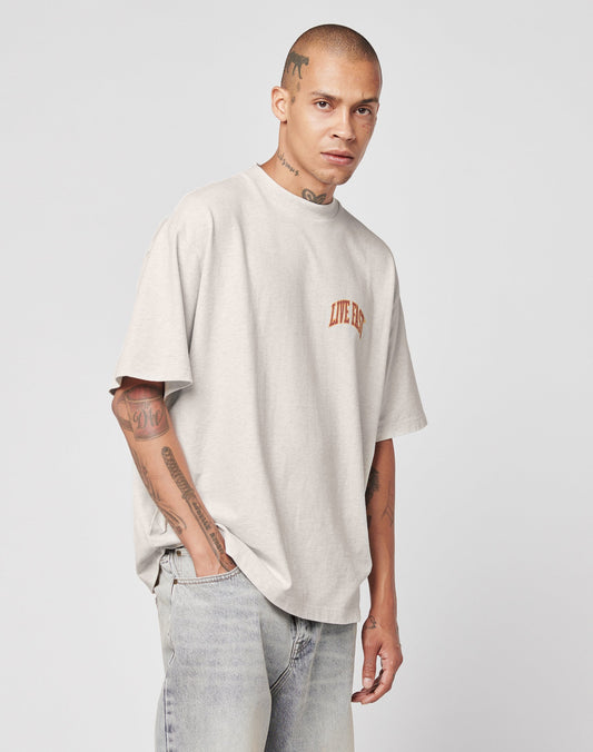 A tattooed man wears the Live Fast Amsterdam Tee by LIVE FAST DIE YOUNG, a vintage-inspired, oversize light gray Baumwolle shirt with brown LIVE BIG lettering, styled with faded blue jeans against a plain light background.