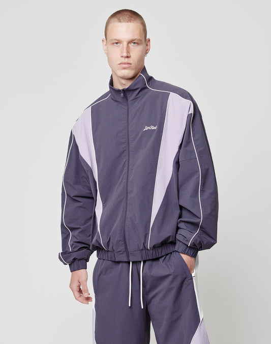 A man with short hair wears the LIVE FAST DIE YOUNG Basic Nylon Trackjacket, a regular fit in purple and lavender with white piping, side slit pockets, and Stay Bold embroidered on the chest. He stands against a plain light background.
