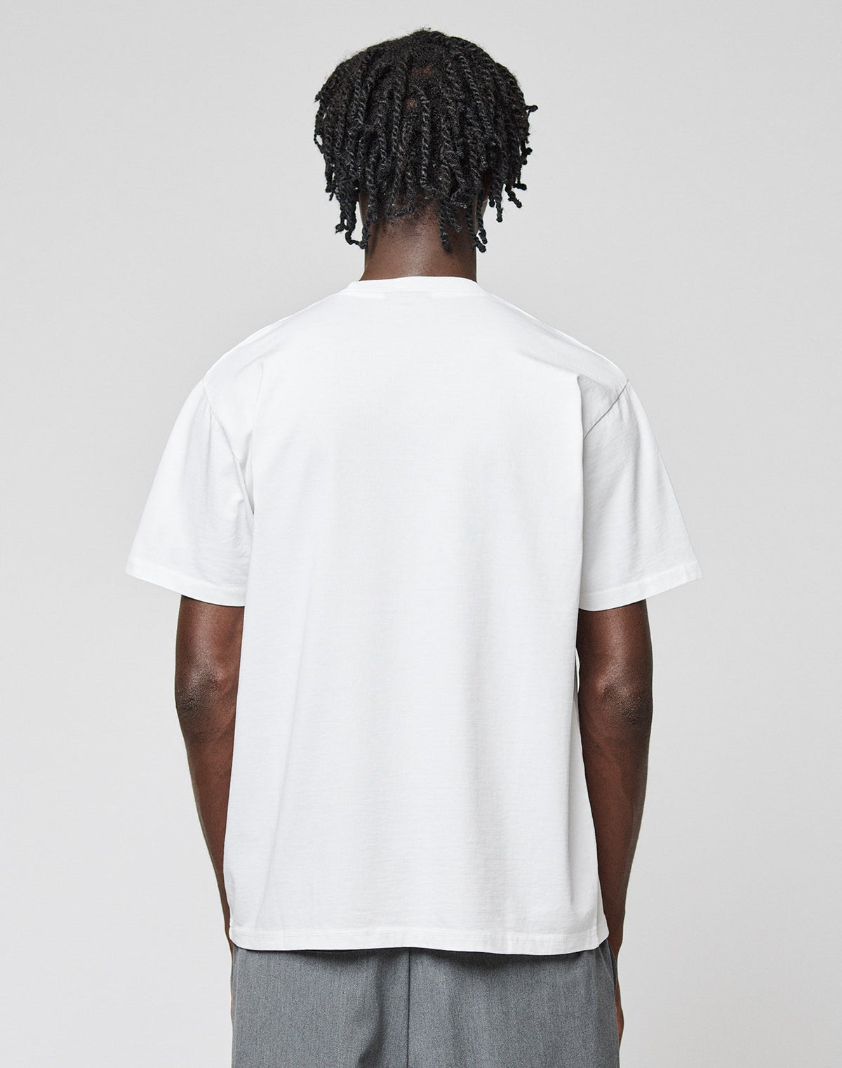 A person with short, twisted hair stands with their back to the camera, wearing the LIVE FAST DIE YOUNG Live Fast Plain Tee in white and light gray pants against a plain light gray background.