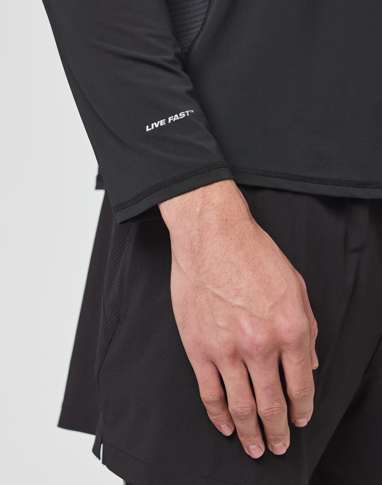 A close-up of a hand resting by their side, wearing the LIVE FAST DIE YOUNG Live Fast Performance Longsleeve with LIVE FAST text on the sleeve, mesh inserts, and black athletic shorts.