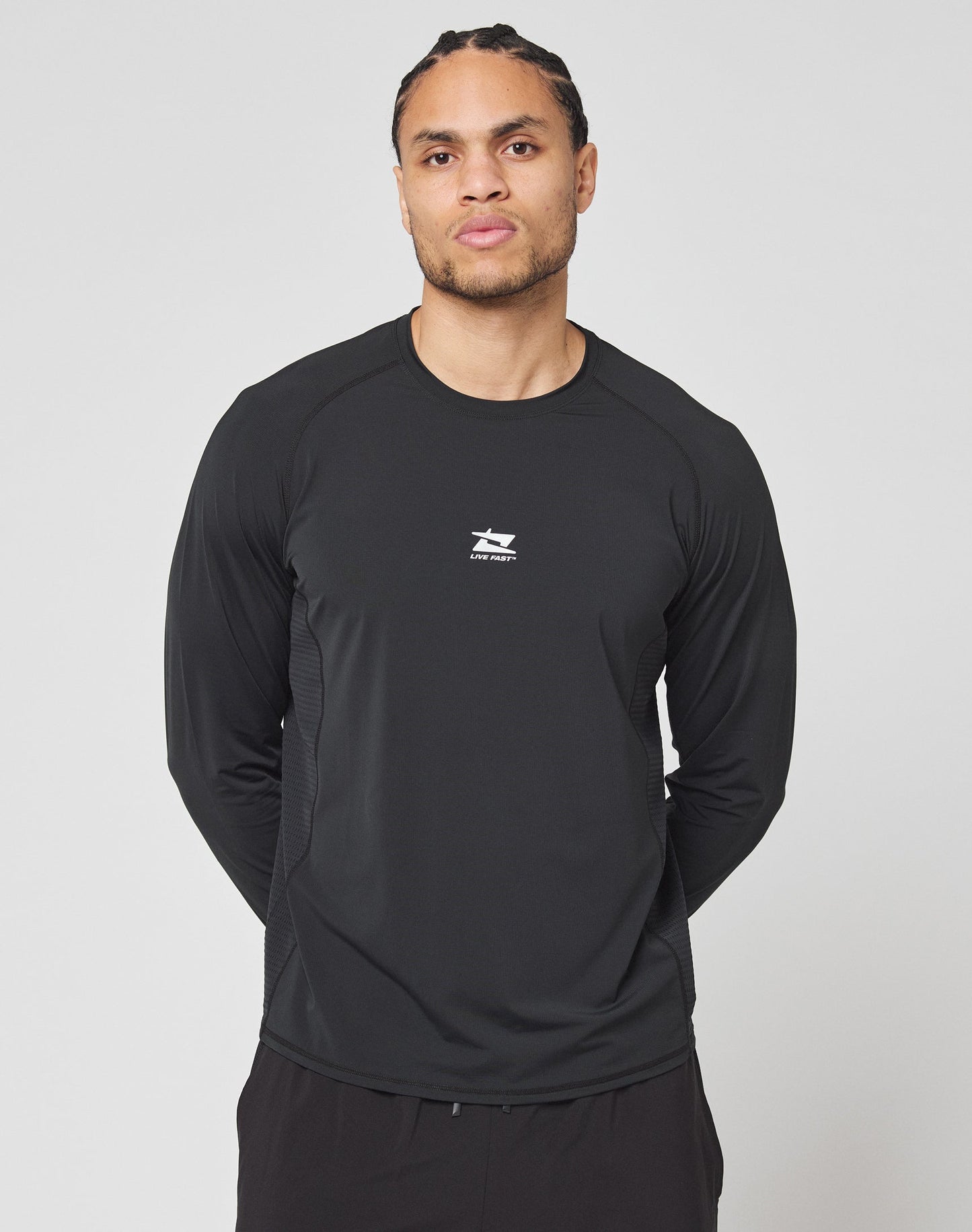 A person with braided hair wears the LIVE FAST DIE YOUNG Live Fast Performance Longsleeve, a black slim fit athletic shirt with a mesh insert and small white logo on the chest, standing before a plain light gray background.
