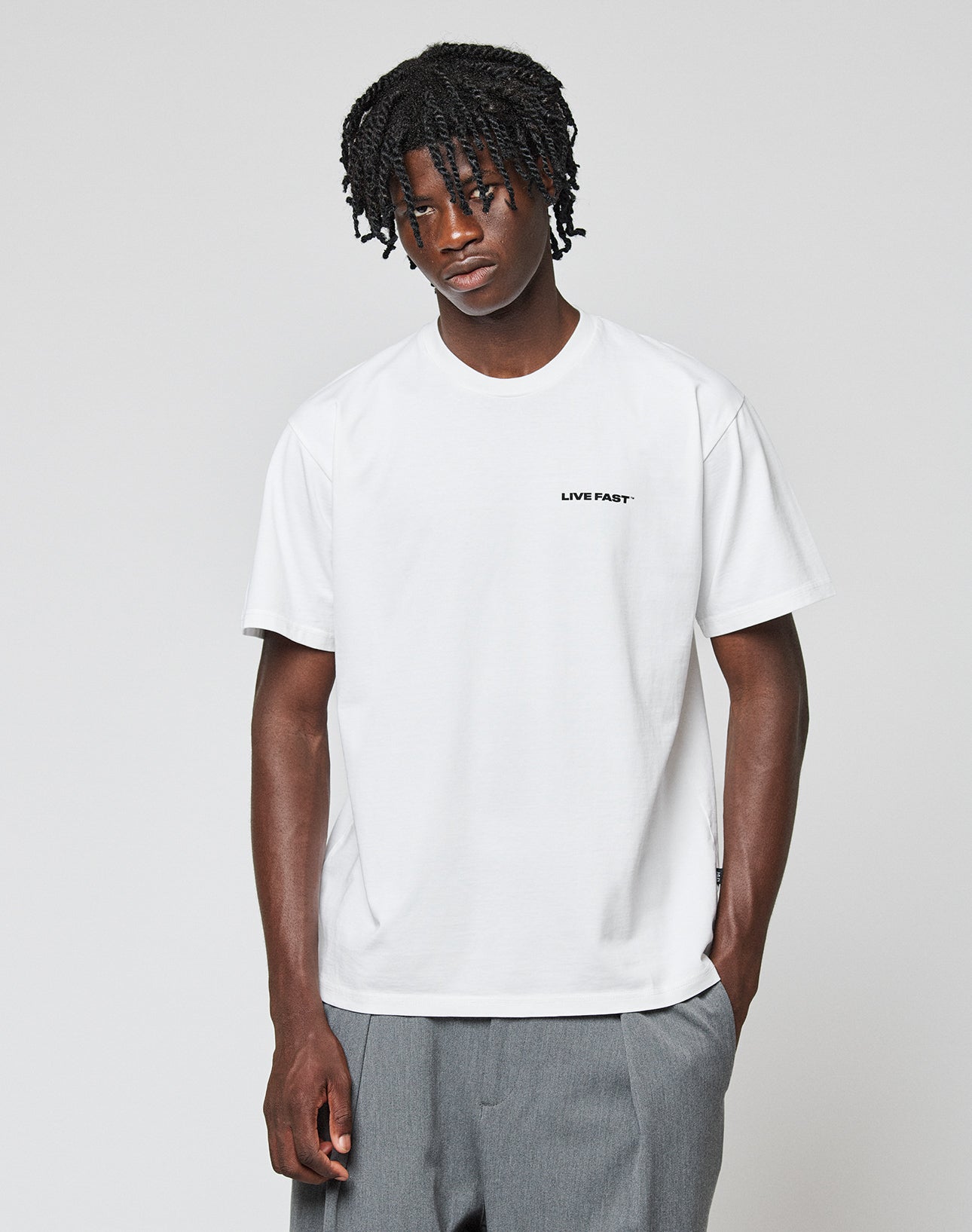 A young man with short, twisty black hair wears the LIVE FAST DIE YOUNG Live Fast Plain Tee (white, regular fit) and loose gray pants. He stands against a plain white background, looking slightly downward with one hand in his pocket.