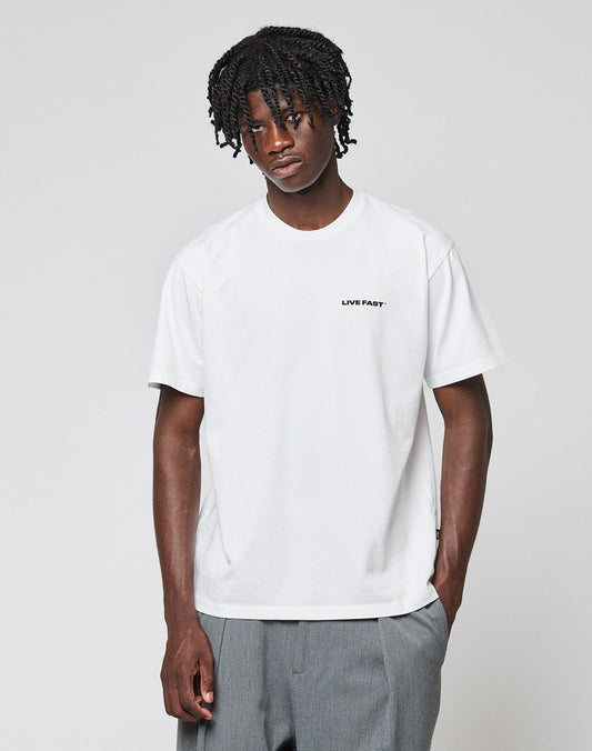 A young man with short, twisty black hair wears the LIVE FAST DIE YOUNG Live Fast Plain Tee (white, regular fit) and loose gray pants. He stands against a plain white background, looking slightly downward with one hand in his pocket.