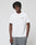 A young man with short, twisty black hair wears the LIVE FAST DIE YOUNG Live Fast Plain Tee (white, regular fit) and loose gray pants. He stands against a plain white background, looking slightly downward with one hand in his pocket.