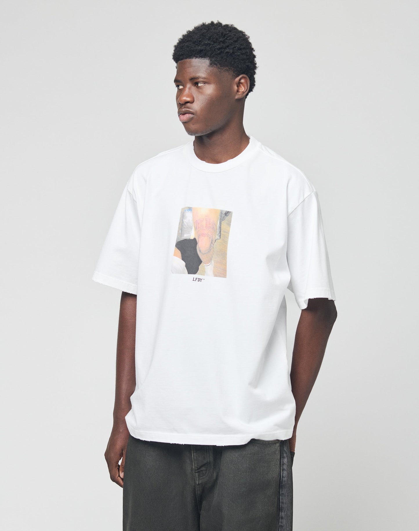 A young man wears the LIVE FAST DIE YOUNG LFDY Sharkhunter Tee—an oversize white t-shirt with a colorful graphic—standing against a plain background, gazing to the side with his hands in his dark pants pockets.