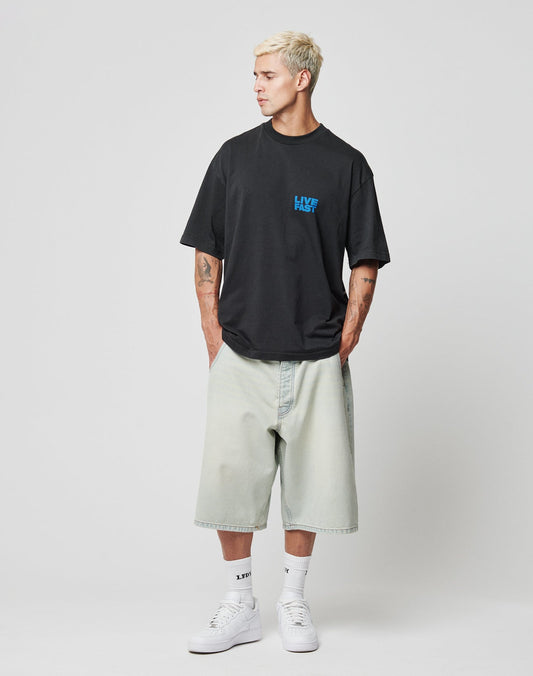 A man with short blond hair wears a loose black T-shirt, LIVE FAST DIE YOUNG LFDY Jorts with Gesäßtasche, white socks, and white sneakers. He stands hands in pockets, looking to the side against a plain white background.