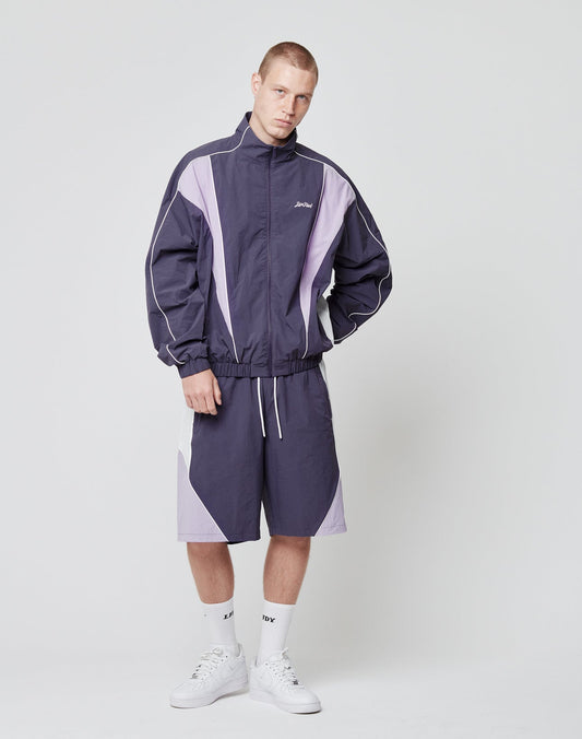A person stands against a plain background wearing the LIVE FAST DIE YOUNG Basic Nylon Trackjacket in dark and light purple, matching shorts with side pockets, white crew socks, and white sneakers. Hands are in their pockets as they look straight ahead.
