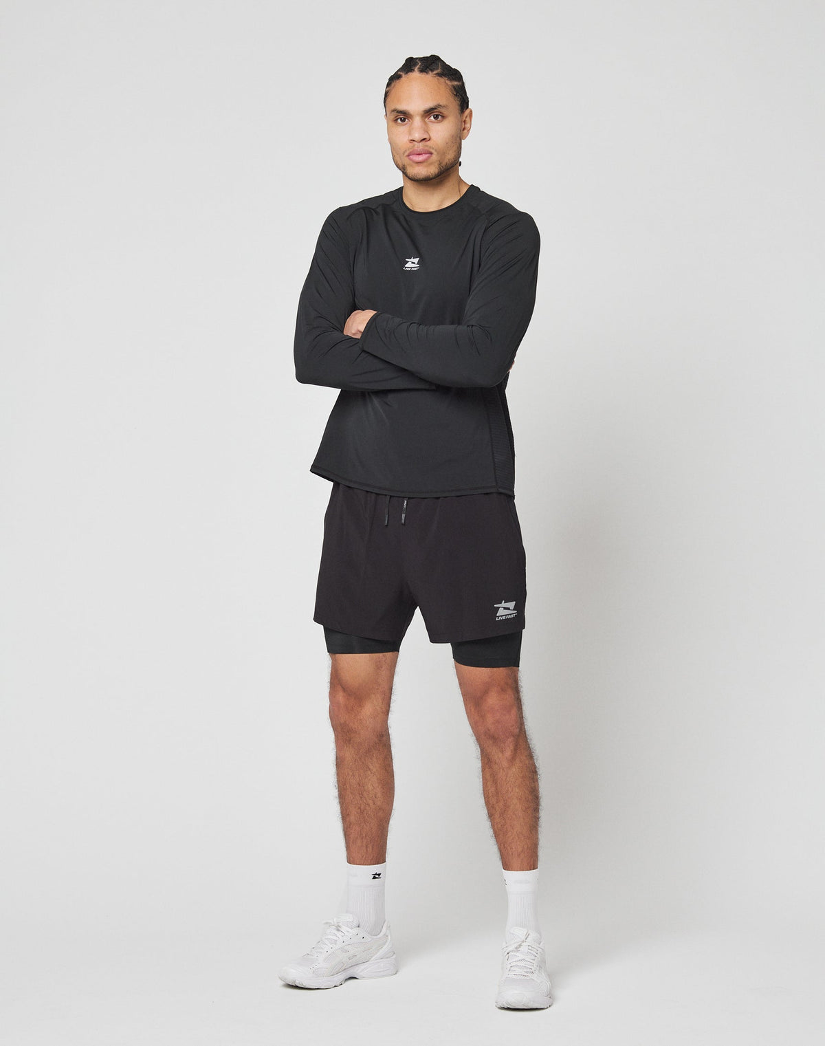 A man stands against a white background wearing the LIVE FAST DIE YOUNG Live Fast Performance Longsleeve, paired with black shorts, white socks, and white sneakers. His arms are crossed as he looks slightly to the side.