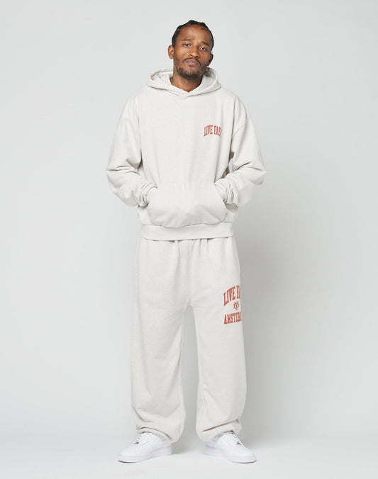 A man stands against a plain background wearing the LIVE FAST DIE YOUNG Live Fast Amsterdam Loose Jogger and matching light gray hooded sweatshirt with red text, paired with white sneakers. His hands are in the sweatshirt pocket as he looks at the camera.