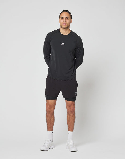 A person stands against a plain white background, wearing LIVE FAST DIE YOUNGs black Live Fast Performance Running Shorts with integrated tights, a black long-sleeve athletic shirt, white socks, and white sneakers. Hands behind back, facing forward.
