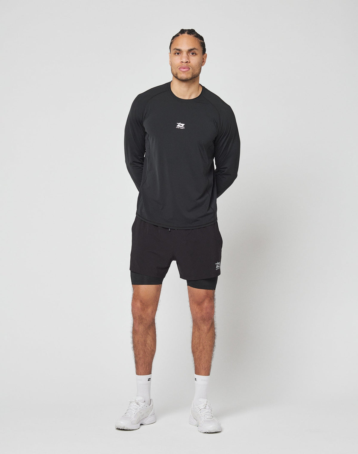 A person stands against a plain white background, wearing LIVE FAST DIE YOUNGs black Live Fast Performance Running Shorts with integrated tights, a black long-sleeve athletic shirt, white socks, and white sneakers. Hands behind back, facing forward.