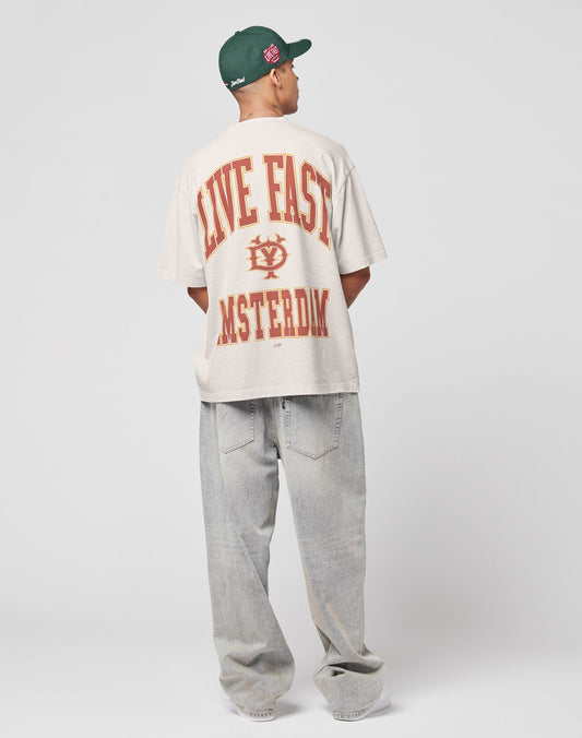 A person in a green cap, baggy light-wash jeans, and the LIVE FAST DIE YOUNG Live Fast Amsterdam Tee—a vintage oversized beige Baumwolle shirt with red LIVE FAST AMSTERDAM text on the back—faces away against a plain gray background.