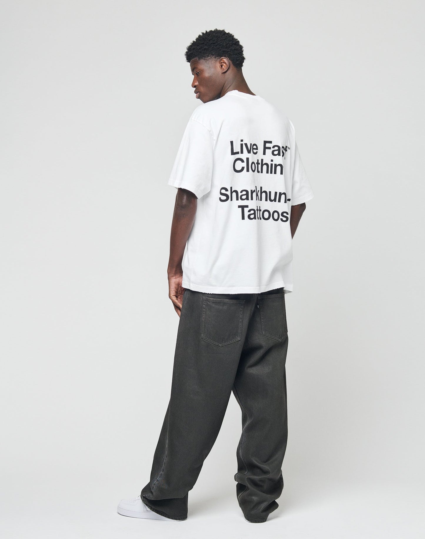 A man stands facing away in the LFDY Sharkhunter Tee by LIVE FAST DIE YOUNG, an oversize white T-shirt with back text, paired with loose dark jeans and white sneakers against a plain light background.