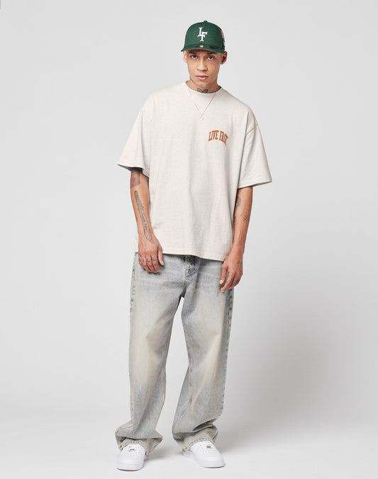 A person in a green cap, beige oversized Live Fast T-shirt, and white sneakers wears LIVE FAST DIE YOUNGs LFDY Baggy Denim—light blue, baggy fit jeans with wide straight legs and a subtle flag label—against a plain gray background.