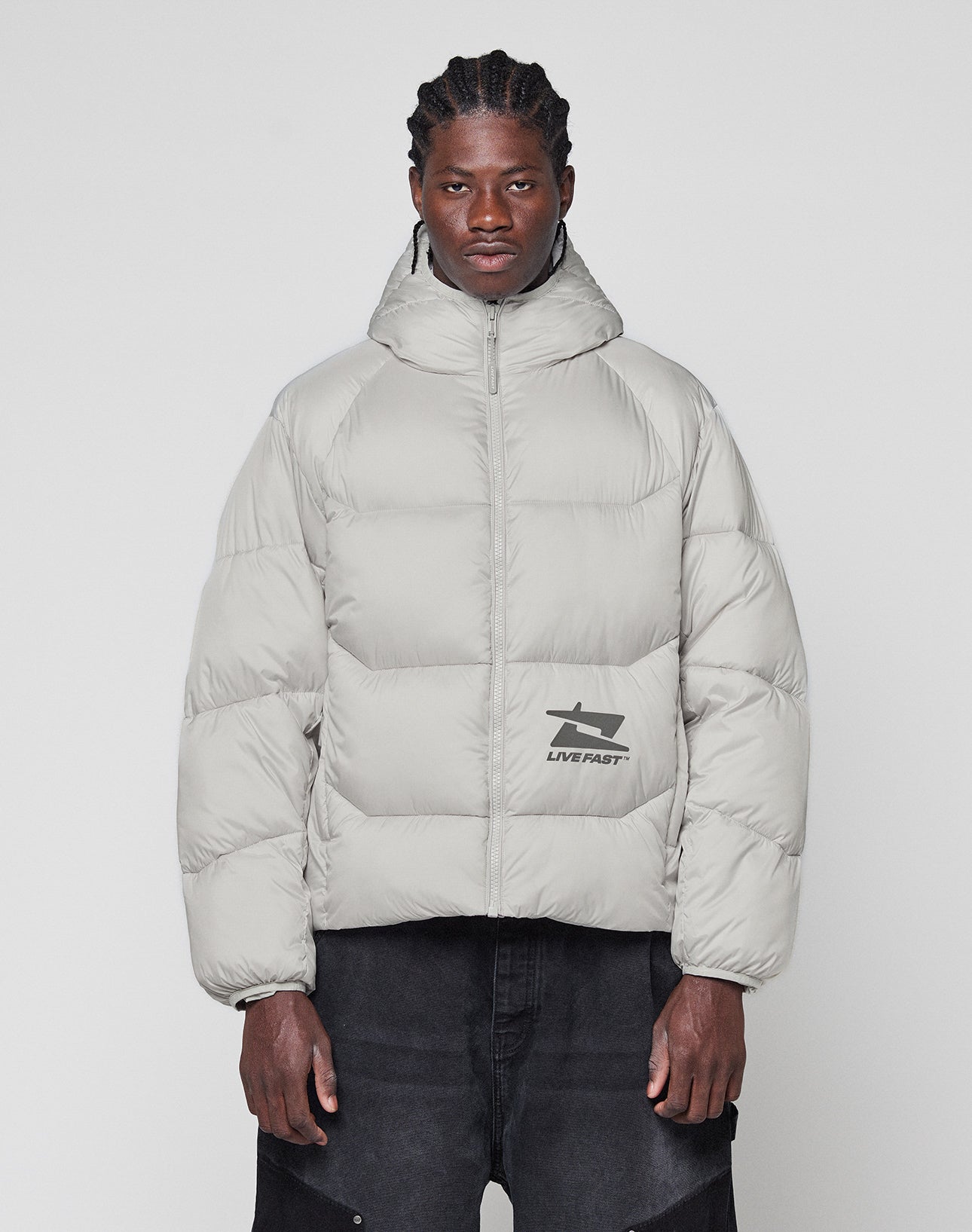 A man with braided hair wears the LIVE FAST DIE YOUNG Live Fast Puffer Jacket, a light gray, oversized, hooded style featuring a reflektierender Print and LIVE FAST graphic, standing against a plain white background.