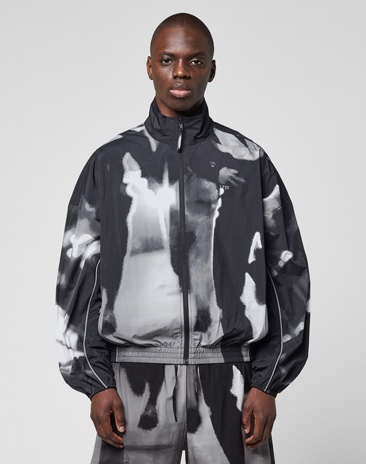 A person wearing a loose-fitting, black and white LFDY Performance Jacket by LIVE FAST DIE YOUNG with a zip-up high collar and matching pants featuring a reflective logo print stands facing forward against a plain light background. | model