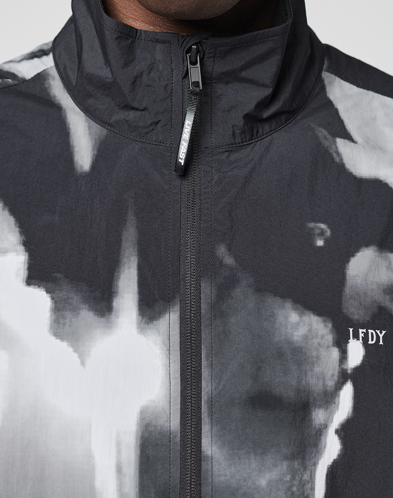 A close-up of a person wearing the LFDY Performance Jacket by LIVE FAST DIE YOUNG, featuring a black and white abstract allover print, high collar, zipper, and reflective LFDY logo on the left chest.