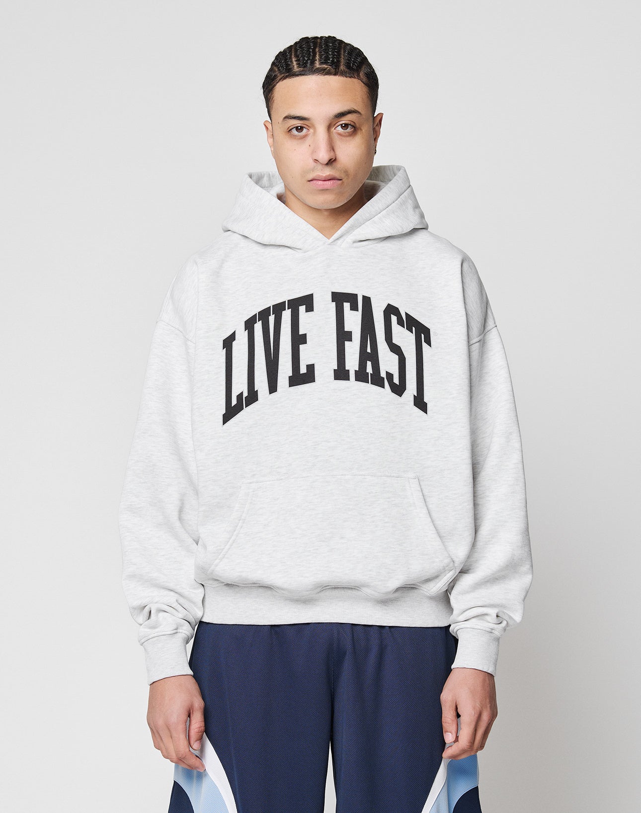 A person with braided hair wears a LIVE FAST DIE YOUNG Live Fast College Hooded in oversize light gray, featuring bold black LIVE FAST text, paired with dark blue athletic pants, standing against a plain white background.