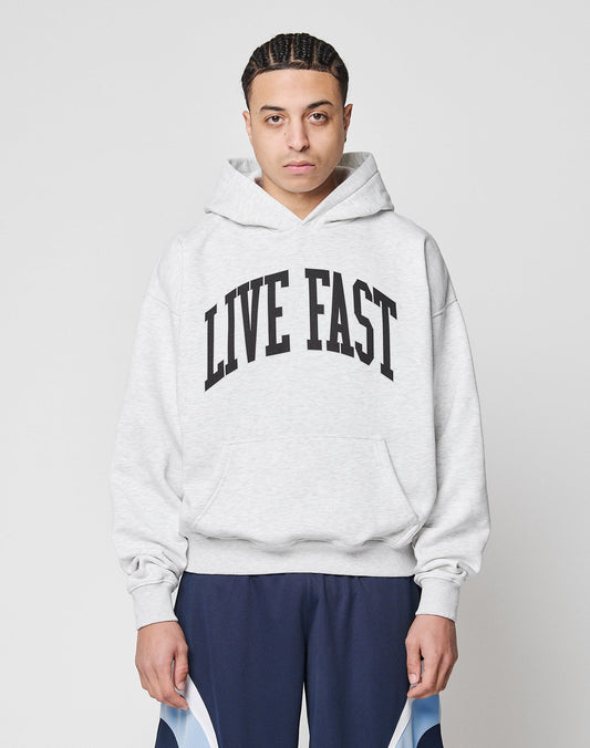 A person with braided hair wears a LIVE FAST DIE YOUNG Live Fast College Hooded in oversize light gray, featuring bold black LIVE FAST text, paired with dark blue athletic pants, standing against a plain white background.