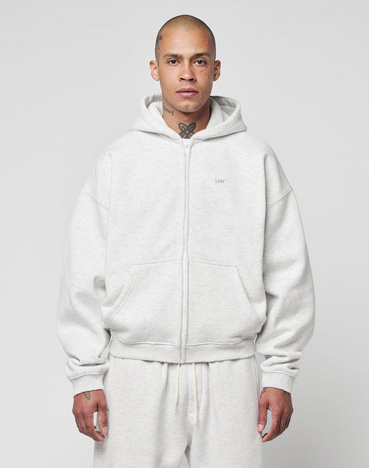 A person with a shaved head and visible tattoos wears the LIVE FAST DIE YOUNG Basic Hooded Zip in light gray with matching sweatpants, standing against a plain light gray background.