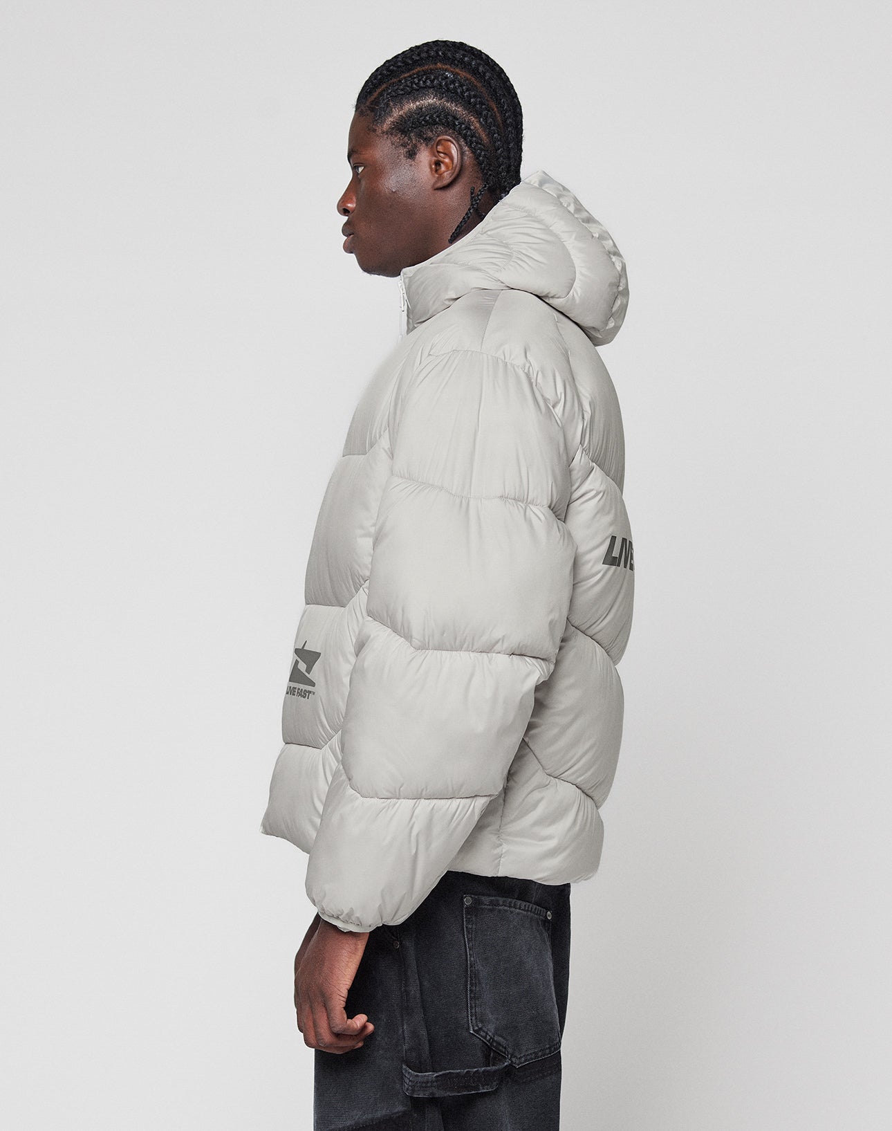 A person with braided hair wears the LIVE FAST DIE YOUNG Live Fast Puffer Jacket in light gray with an oversize fit, hood, reflektierender Print, and subtle sleeve and side graphics, standing in profile against a plain white background.
